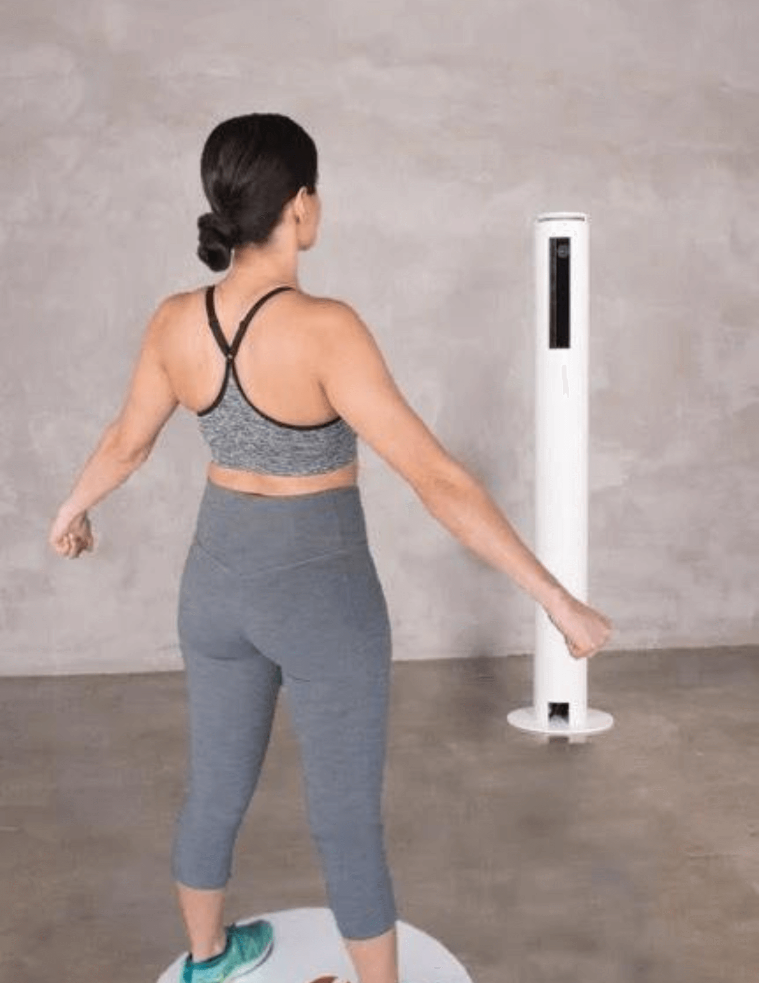 A woman stands on a platform with her arms outstretched, being scanned by a 3D body scanner. The scanner is a white cylinder with a black panel and is positioned in front of a concrete wall.